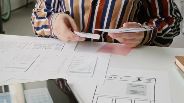 Medium midsection shot of anonymous young female mobile application developer working with wireframe prototype cutouts at desk, arranging in sequence on paper, tablet and smartphone nearby