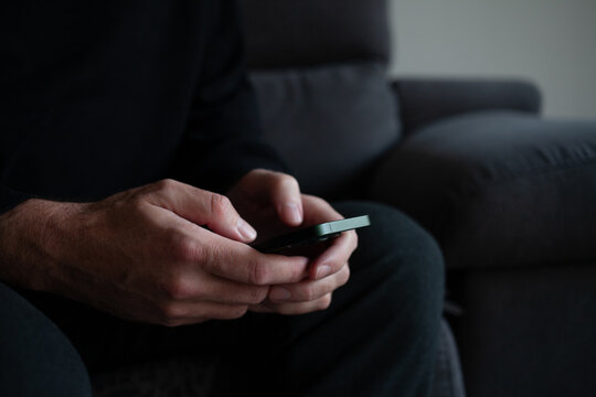 Man using smartphone while sitting on a sofa at home, close-up of hands holding mobile phone in soft natural light. Modern lifestyle, communication, online browsing and digital technology concept
