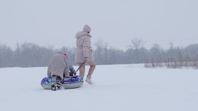 Mother pulls a colorful sled with kids in winter clothes across a snowy field. Snow falls gently as they enjoy their time outside together