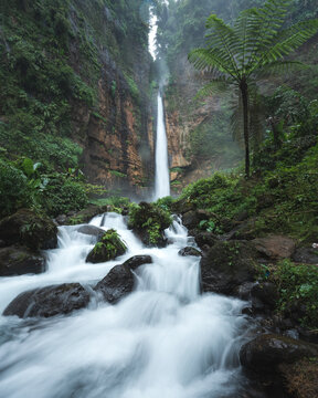 Aerial view of a powerful waterfall cascading down moss-covered cliffs into a rocky riverbed, surrounded by lush green forest, Probolinggo, Jawa Timur, Indonesia.