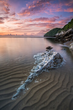 View of the sunrise paints the sky in hues of pink and purple, reflecting on the serene water, with a rock sitting on the beach, Hua Hin, Phetchaburi, Thailand.