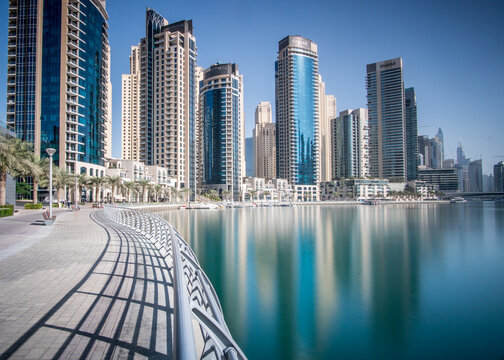 View of gleaming skyscrapers reflect in tranquil waters, palm trees sway, casting shadows on the promenade in Financial District, Dubai, United Arab Emirates.