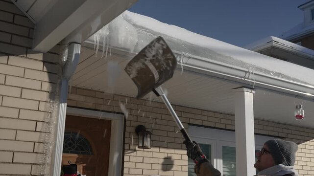 Homeowner uses a shovel to knock large icicles from a snow-covered roof gutter on a sunny winter day, highlighting seasonal home maintenance and ice hazard removal. 15032