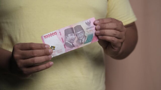 Close-up of a man holding an Indonesian rupiah note, showing cash payment, personal finance, budgeting, and everyday spending concept indoors.