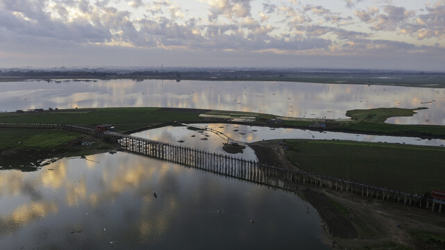 Aerial view of the U Bein Bridge stretching across the serene Taungthaman Lake, its reflection shimmering in the water under a soft, cloudy sky, Mandalay, Mandalay Region, Myanmar.