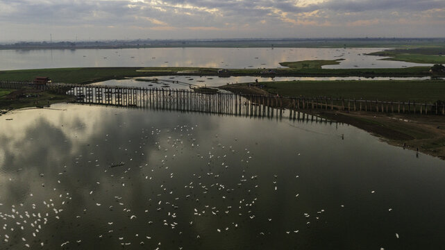 Aerial view of U Bein Bridge stretching across the tranquil Taungthaman Lake where birds gather under a serene sky, Mandalay, Mandalay Region, Myanmar (Burma).