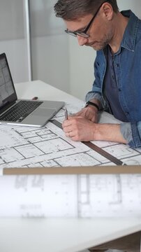 Architect sketching building plans on paper using a ruler and pen while working digitally on a laptop with a blueprint software displaying a floor plan