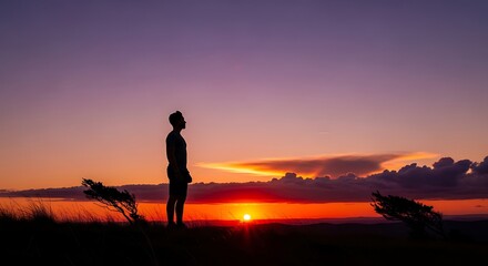 Silhouetted figure stands on a grassy hill watching the vibrant sunset.
