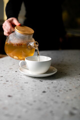 A person is pouring hot herbal or fruit tea from a transparent glass teapot with a wooden lid into a white ceramic cup on a saucer, set on a light granite countertop