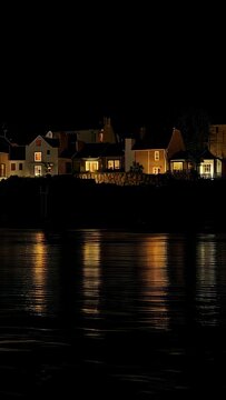 Vertical video: Rippling water reflecting lit houses at shore as lights shimmering along stone wall