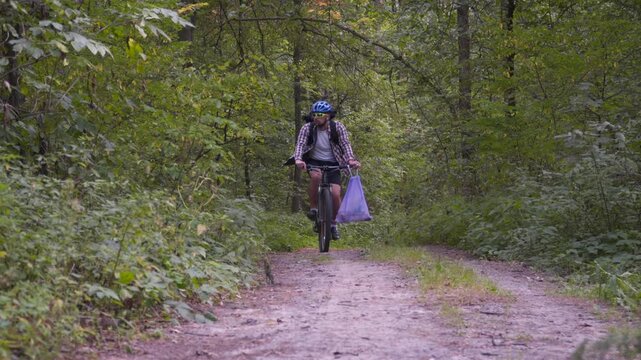 Cyclist stops on a forest trail, shows a plastic bag full of collected trash, and continues biking. Concept of plogging, environmental cleanup, nature care, and sustainable sport