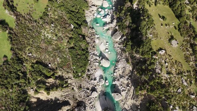 A mesmerizing top-down shot of a pure turquoise stream flowing through grey boulders and lush green vegetation. The contrast between the vibrant water color and the rugged stones creates a natural abs