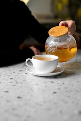 A close-up shot of an unrecognizable person pouring hot herbal tea from a glass teapot with a wooden lid into a white ceramic cup and saucer on a speckled countertop.