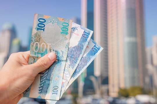 Hand holding currency, Qatari riyal banknotes, with modern skyscrapers in Doha city creating a financial wealth and business concept