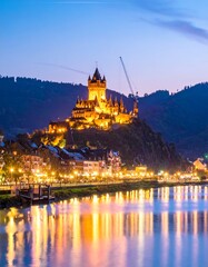 Obraz premium Illuminated medieval castle atop a hill at dusk. A river reflects city lights and mountain slopes on either side