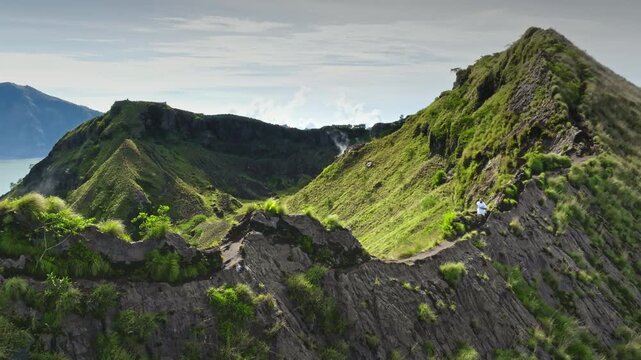 Mount Batur, Bali: Man active hiking narrow green mountain ridge connecting volcanic peaks, enjoy the challenge adventure with crater steaming in background and a vast landscape. Aerial drone flight