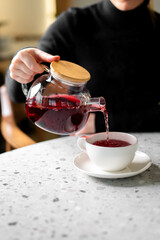 A person in a black sweater pours vibrant red fruit tea from a glass teapot with a wooden lid into a white ceramic cup and saucer on a modern speckled table in a cafe setting
