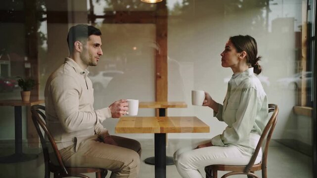Charming young couple sitting at a wooden table in a cozy coffee shop, drinking from white mugs and engaging in a pleasant conversation, seen through the cafe window with reflections