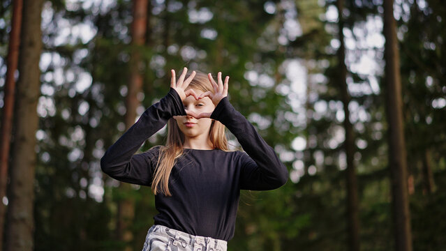 Teen girl gen Alpha stands outdoors in park, making a heart shape with her hands, looking at camera, smiling gently and radiating love, positivity.
