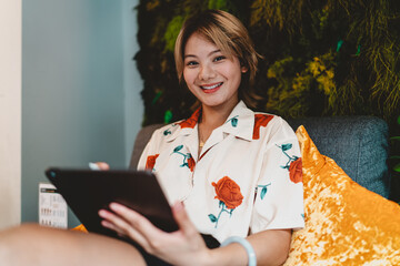 Asian woman smiling warmly while holding tablet on sofa, representing balance, technology-driven creativity, and freelance independence in digital workspace. © BullRun