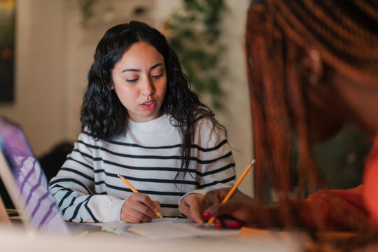 Two young women are studying together at a table, writing with pencils on papers, with a laptop nearby.