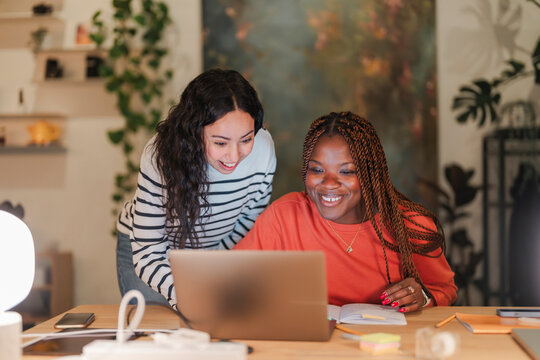 Two women collaborate on a project, smiling as they look at a laptop screen in a cozy, well-lit room.