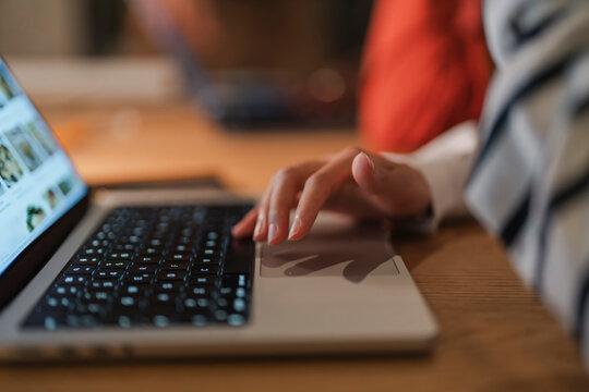 A person's hand is shown using a laptop's touchpad, with the screen displaying images. The scene is set on a wooden desk.