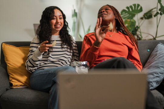 Two young women laugh while watching something on a laptop and eating snacks on a couch.
