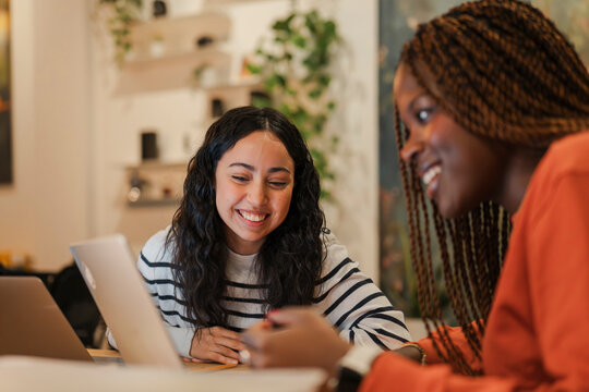Two young women are smiling and interacting while working on laptops in a bright, modern cafe setting.