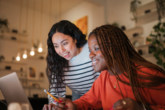 Two young women collaborate on a project, smiling as they look at a laptop screen together in a cozy, dimly lit room.