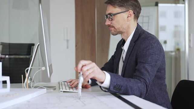 Male architect working on architectural blueprints, using a compass next to a computer while developing new building designs. Business and architecture concept