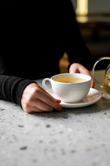 A close-up shot of hands in black sleeves holding a white cup of hot herbal tea on a saucer, resting on a gray granite counter
