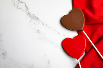 Heart-shaped chocolate candies arranged on rich red fabric over a white marble background with ample copy space. 
