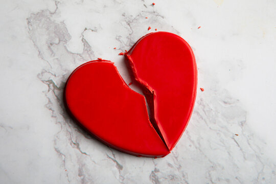 A broken red candy heart split into pieces on a white marble background, symbolizing heartbreak, breakup, and emotional pain. The clean, minimal composition makes this image ideal for relationship art