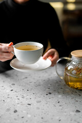A close-up, vertical shot of a person in a black sweater holding a white cup and saucer filled with hot, yellow herbal tea, with a glass teapot visible nearby on a white speckled counter.