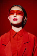 Young fashionable woman in bright red blazer and shirt looking confidently at the camera. She wears oversized red translucent sunglasses. Studio portrait with vivid red background, creative beauty