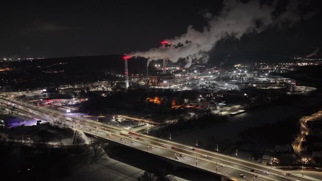 Zoomed-in nighttime drone shot of an industrial facility in Vilnius, Lithuania, with tall chimneys emitting large plumes of steam, illuminated by red warning lights over a snowy landscape.