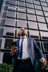 Fototapeta premium Low-angle view of businessman standing outside modern building, looking up while holding smartphone and tablet, symbolizing vision, ambition and connected business mindset.