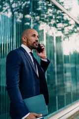 Businessman speaking on phone while holding tablet, standing confidently near glass building,...