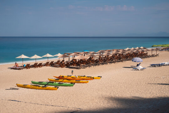 View of golden sand meets turquoise water under a bright sky, with rows of beach umbrellas and colorful kayaks lining the shore, Lefkada, Greece.
