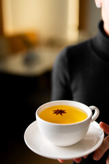 A close-up, horizontal photo of a person in a black sweater holding a white cup and saucer containing golden star anise tea, with a single star anise pod floating in the center.