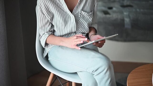 Individual engages with a tablet while seated in a contemporary chair. The setting features a wooden table and simple decor. Natural light fills the room