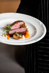 A chef presents a modern plated dish of perfectly cooked sliced duck breast with crispy skin, garnished with pureed vegetables, herbs, and sauce