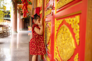 Obraz premium Adorable Little Girl in Traditional Red Cheongsam Standing by Golden Carved Temple Doors,A cute young Asian girl wearing a festive red Chinese dress (Qipao) with gold floral patterns,