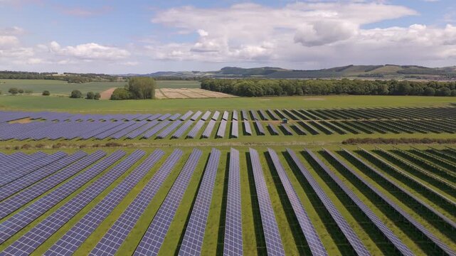 Aerial view of rows of solar panels in a vibrant green field, contrasting with the blue panels under a partly cloudy sky, Perth, Scotland, United Kingdom.
