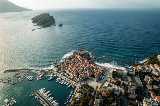 Aerial view of the old town's terracotta roofs contrast with the azure Adriatic Sea, framed by the rugged coastline and Sveti Nikola Island, Budva, Budva Municipality, Montenegro.