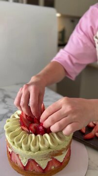 Top view of hands in pink chef shirt and apron placing fresh strawberries and baby basil on luxury fraisier cake. Gourmet dessert finishing touches, professional pastry styling