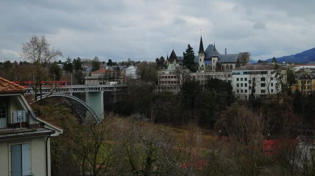 The industrial steel arches of the Kirchenfeldbr&uuml;cke span across the turquoise Aare river, leading to the castle-like architecture of the Bern Historical Museum nestled among winter trees under a dram