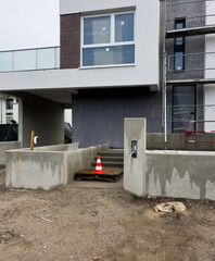 Modern new apartment building under construction concrete walls brick facade large windows balcony scaffolding gray panels entrance stairs traffic cone pallet dirt ground urban.