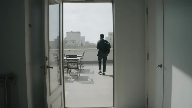 Walking commuter in blazer and backpack through open glass door onto rooftop past table to skyline